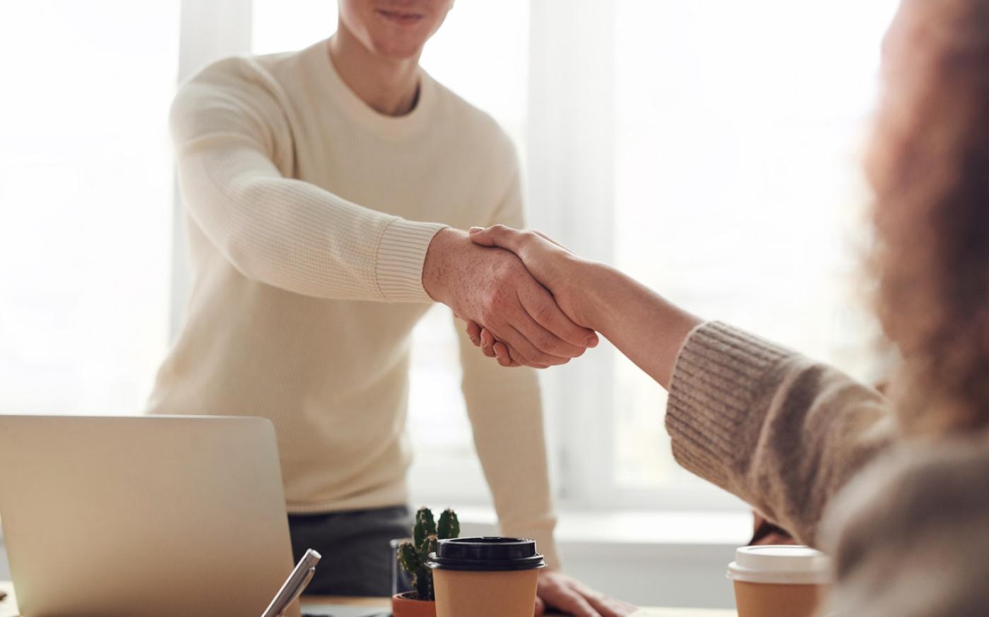 Man and woman shaking hands over a desk. On the desk is a laptop and a coffee cup.