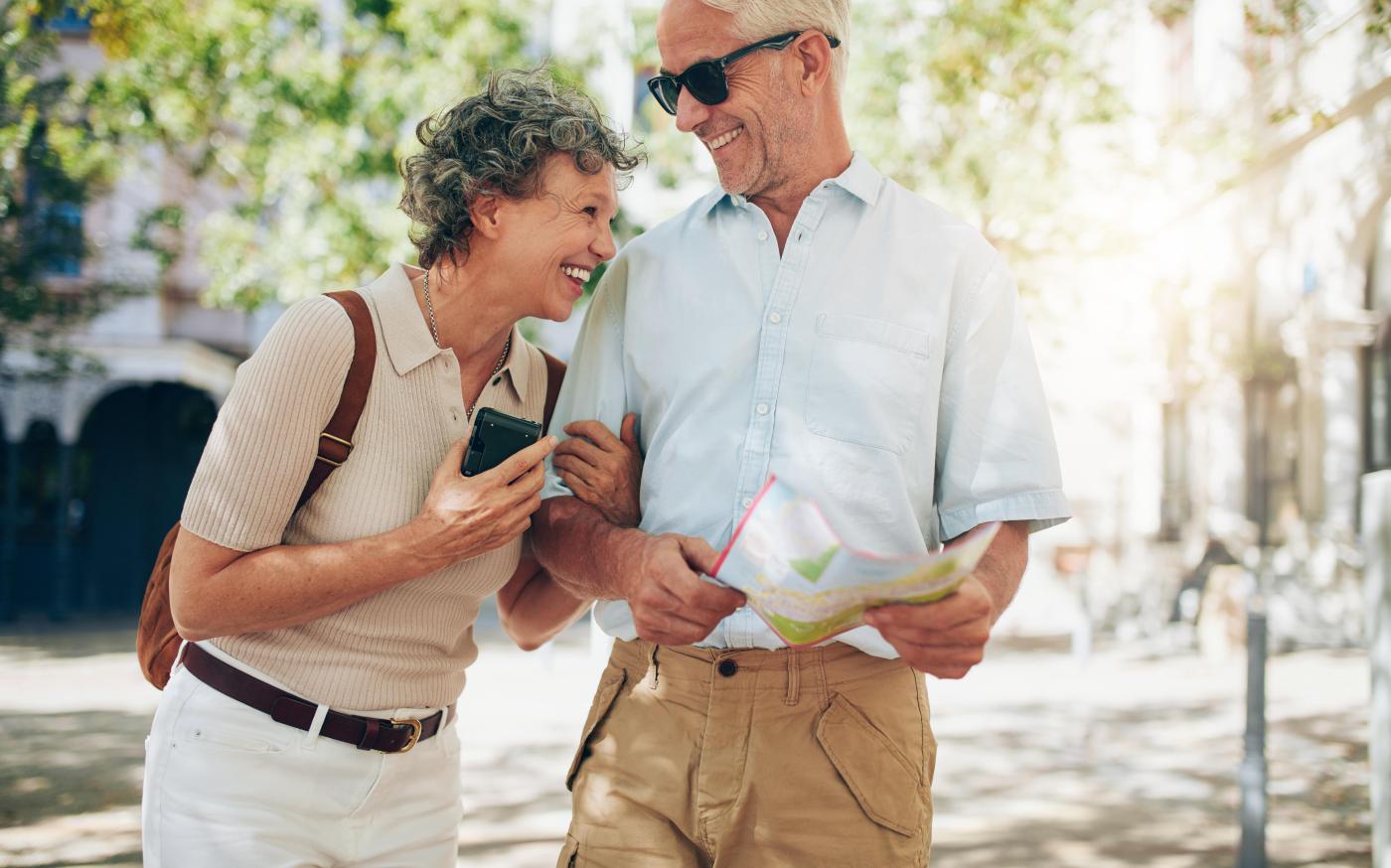 Elderly man and woman laughing. The man is holding a map, and the woman is wearing a backpack. They look like they are travelling.