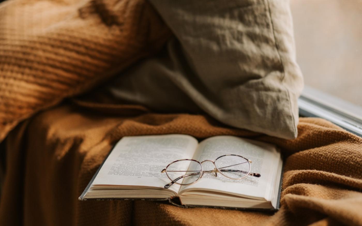 An open book on a windowsill. The book is resting on a knit blanket, and there is a pair of reading glasses on top of the book.